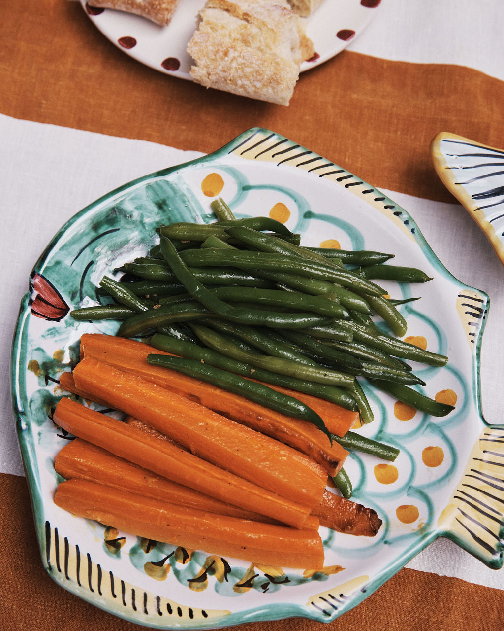 Steamed green beans and carrot sticks are arranged on the Rico Serving Platter, a charming fish-shaped dish, with bread visible in the background.