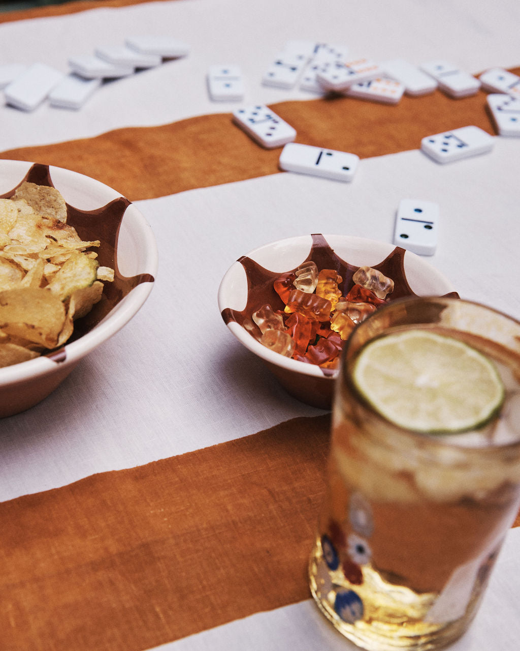 A table with Stella Nesting Bowls filled with potato chips and gummy bears, a glass of iced drink garnished with lime, and dominoes scattered in the background.
