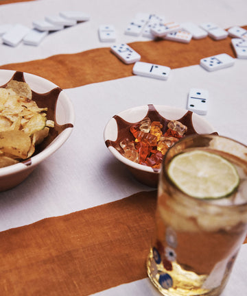 A table with Stella Nesting Bowls filled with potato chips and gummy bears, a glass of iced drink garnished with lime, and dominoes scattered in the background.