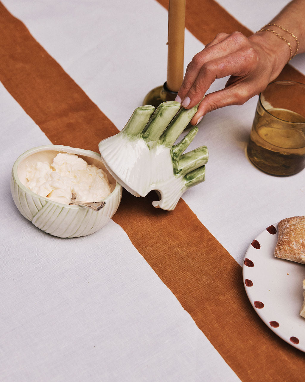 A hand lifts the Fennel Sauce Bowl lid—an Italian ceramic piece—from a bowl of ricotta on a table set with a candle, drink, and bread.