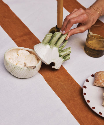 A hand lifts the Fennel Sauce Bowl lid—an Italian ceramic piece—from a bowl of ricotta on a table set with a candle, drink, and bread.