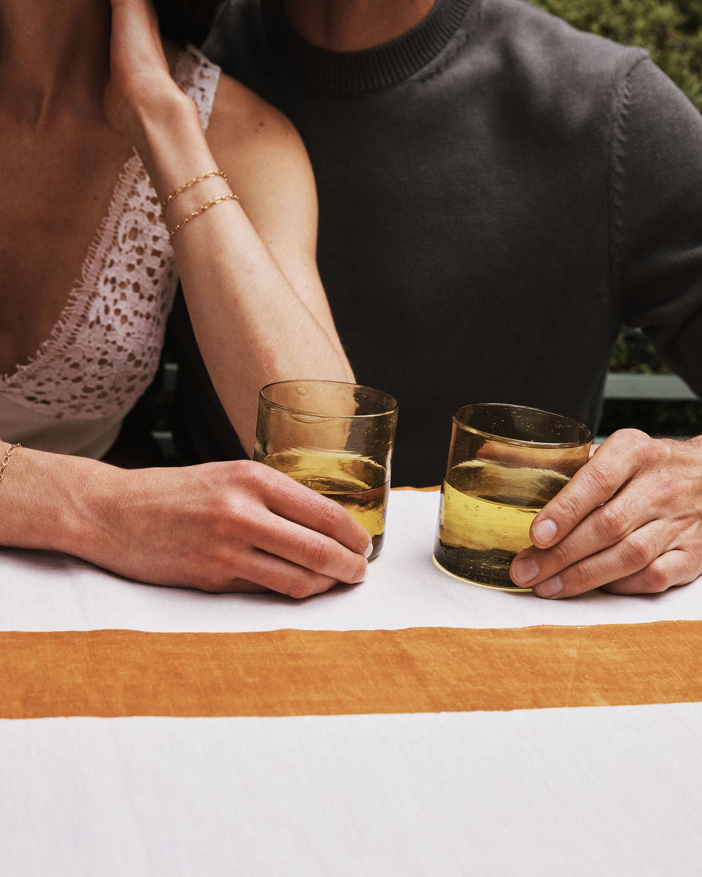 Two people sit side by side at a table, each holding an Elodie Straight Glass filled with a yellowish drink. Their faces are out of frame and their arms rest on the tablecloth, showcasing the elegance of this set of 6 Moroccan glasses.