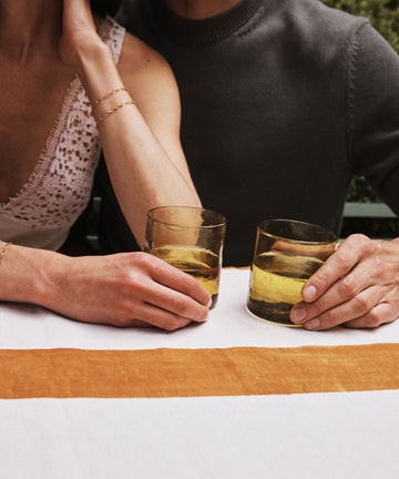 Two people sit side by side at a table, each holding an Elodie Straight Glass filled with a yellowish drink. Their faces are out of frame and their arms rest on the tablecloth, showcasing the elegance of this set of 6 Moroccan glasses.