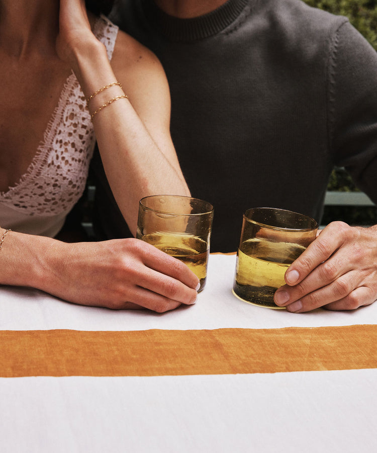 Two people sit side by side at a table, each holding an Elodie Straight Glass filled with a yellowish drink. Their faces are out of frame and their arms rest on the tablecloth, showcasing the elegance of this set of 6 Moroccan glasses.