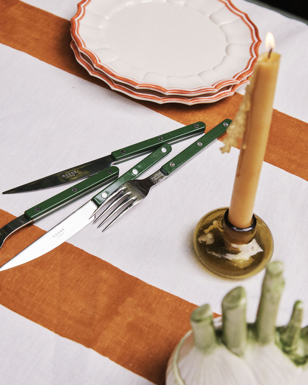 A table set with stacked plates, a striped tablecloth, a Bistrot 5 Piece Cutlery Set with green handles—including knife, fork, and scissors—and a lit candle in a holder.