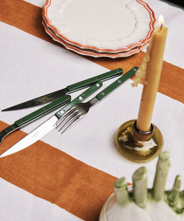 A table set with stacked plates, a striped tablecloth, a Bistrot 5 Piece Cutlery Set with green handles—including knife, fork, and scissors—and a lit candle in a holder.
