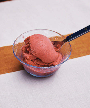 A Lauren Ice Cream Bowl, filled with pink ice cream and a spoon, rests on a white tablecloth featuring a brown stripe.