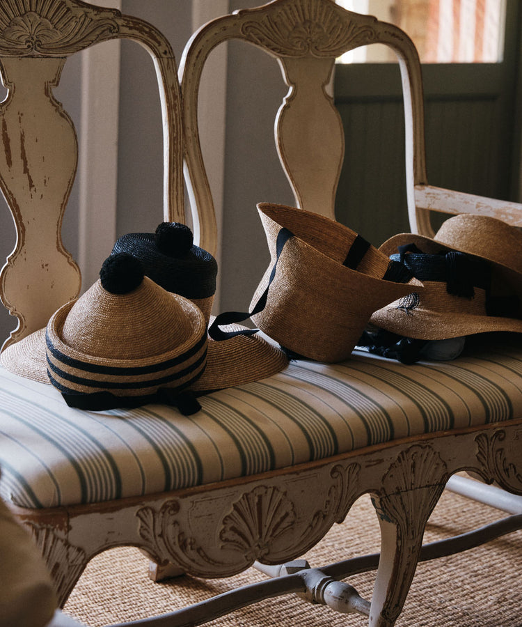 Two Circus Hats and two straw berets with black bands, pom-poms, and long black ties are placed on a vintage upholstered bench featuring intricate wooden details.