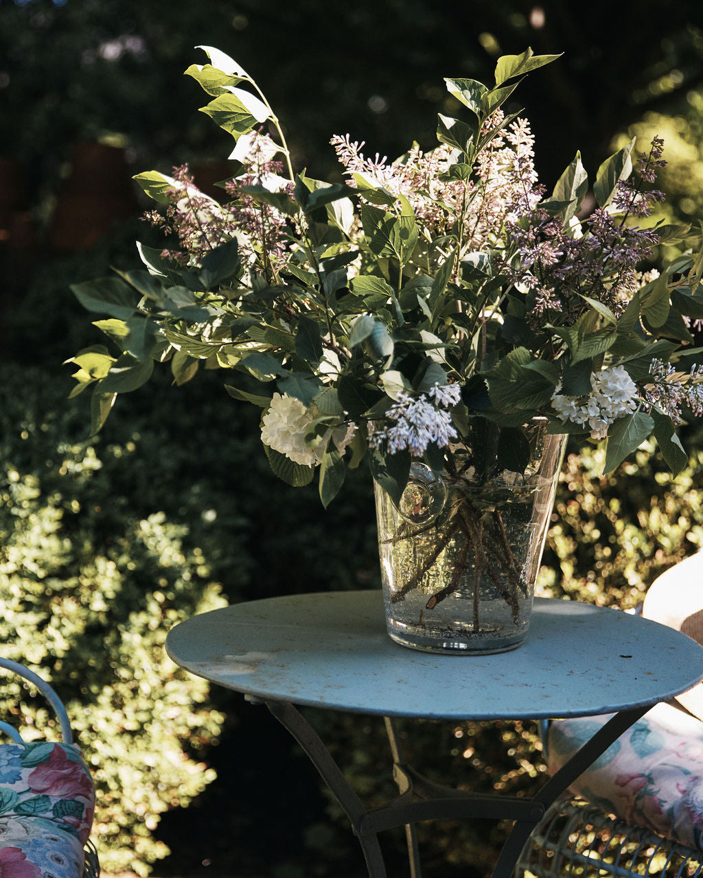 The Lauren Champagne Bucket, filled with assorted leafy branches and flowers, sits on a round metal table outdoors next to a floral cushioned chair.