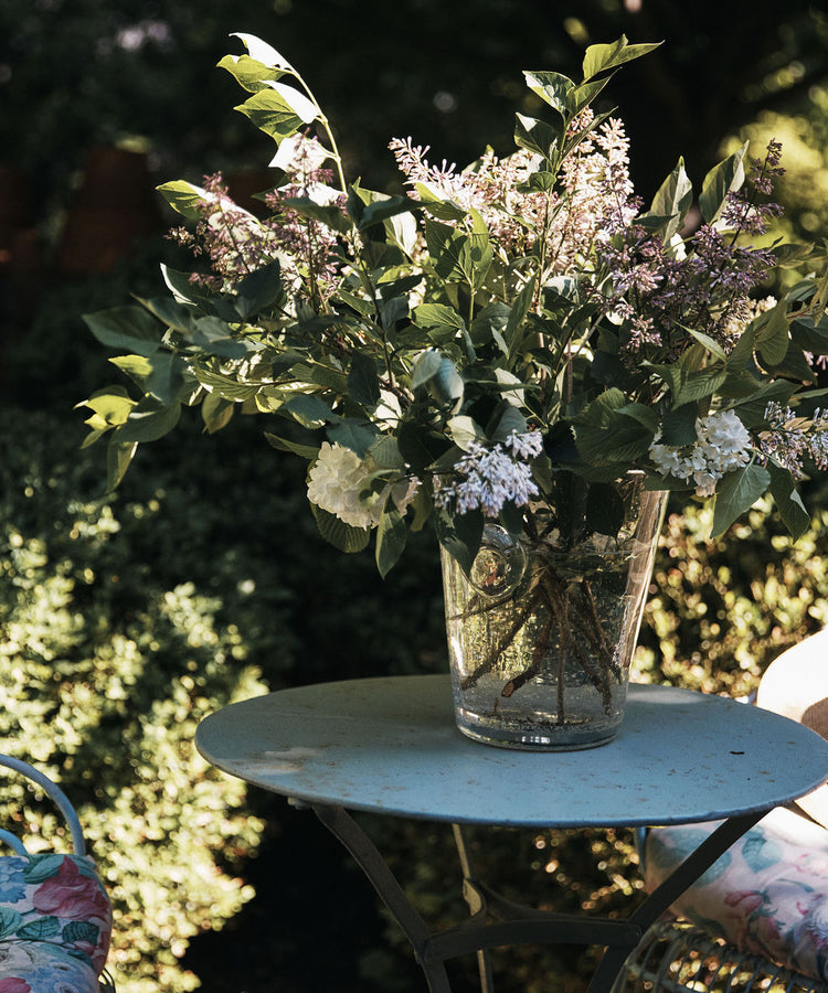 The Lauren Champagne Bucket, filled with assorted leafy branches and flowers, sits on a round metal table outdoors next to a floral cushioned chair.
