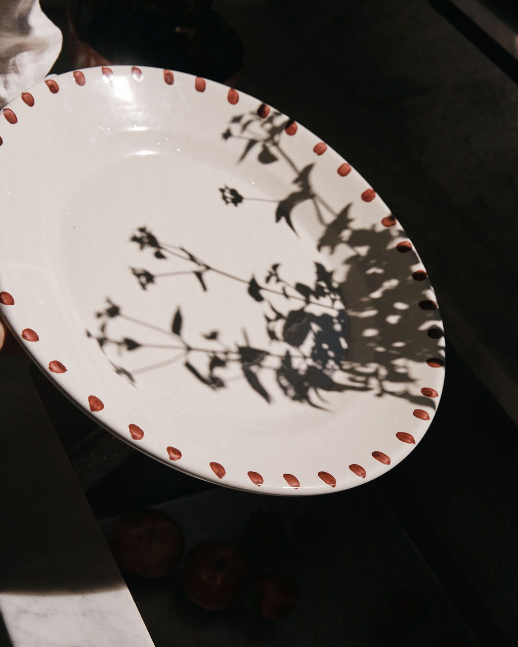 The Nina Serving Plate, a white piece with a dotted red rim, is held up as leafy branch shadows fall across its surface—an elegant dinnerware item from the Nina collection.