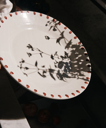 The Nina Serving Plate, a white piece with a dotted red rim, is held up as leafy branch shadows fall across its surface—an elegant dinnerware item from the Nina collection.