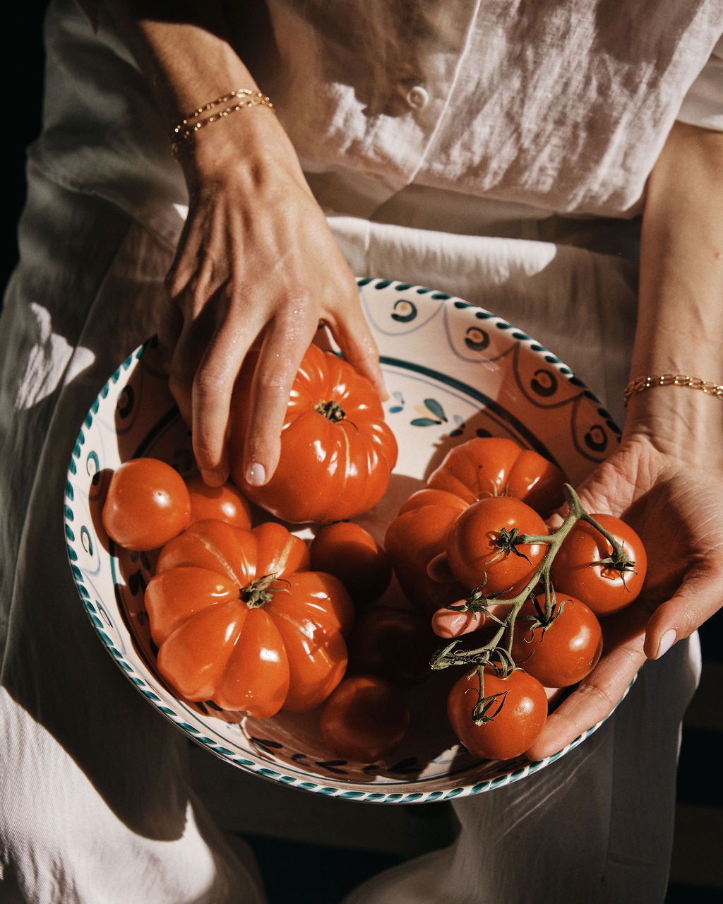 A person in a white outfit holds an Ottoline Serving Bowl filled with various sizes of ripe red tomatoes.