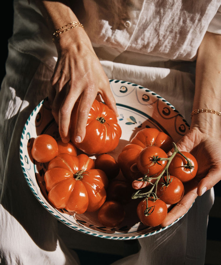 A person in a white outfit holds an Ottoline Serving Bowl filled with various sizes of ripe red tomatoes.