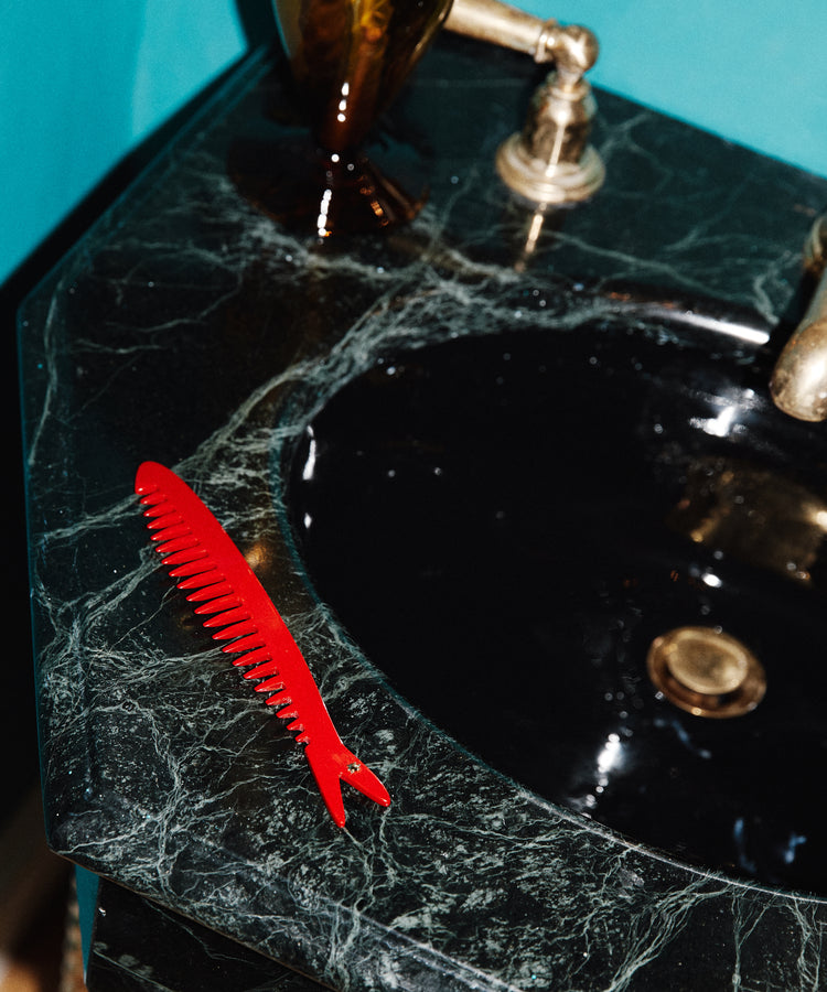 A Capri Sardine Comb rests on a dark green marble bathroom sink next to a brass faucet.