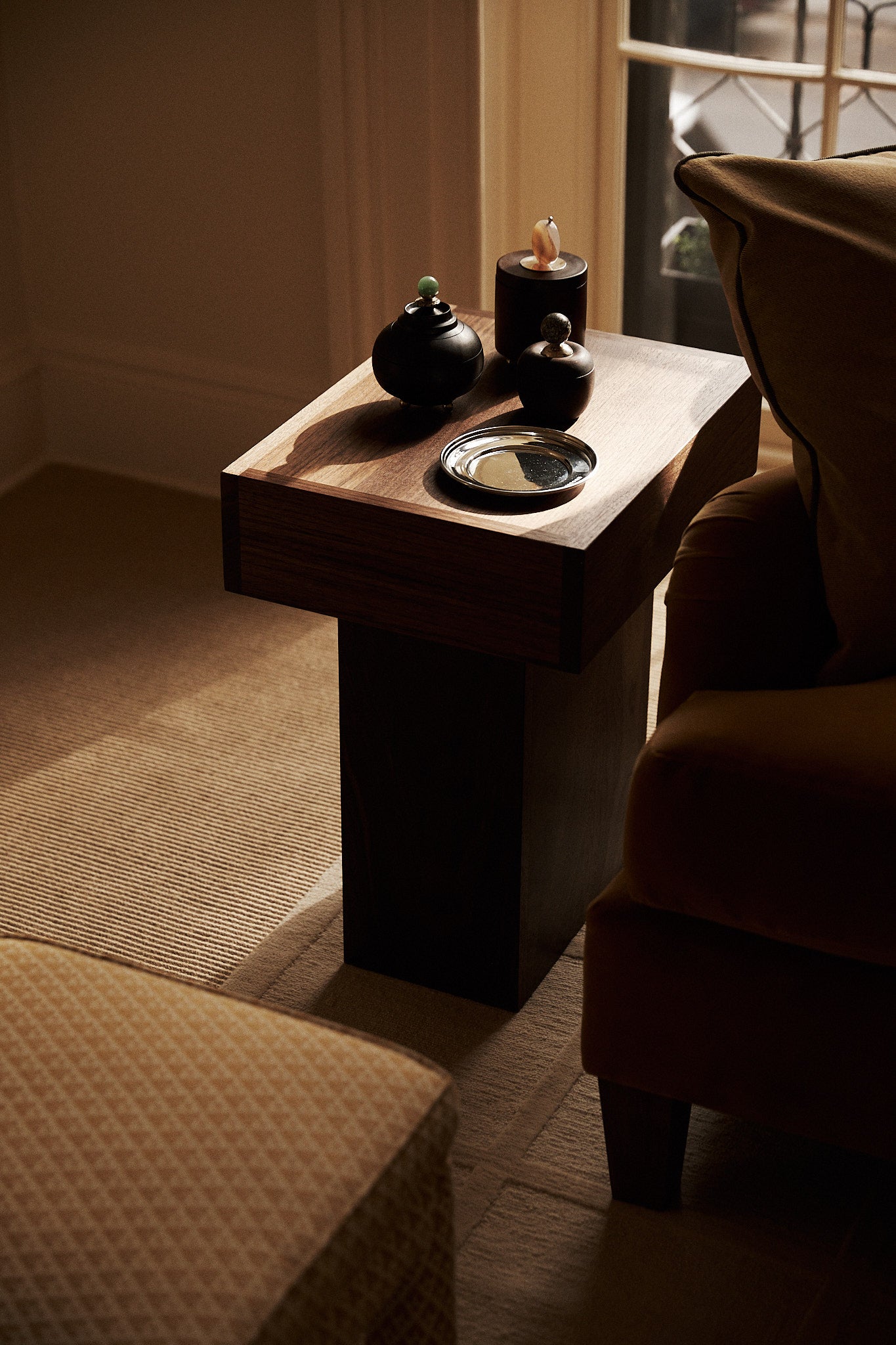 The wooden side table, a timeless essential, holds two decorative jars and the Greta Small Bread Plate as a catchall tray, positioned between an armchair and a window in a softly lit room.