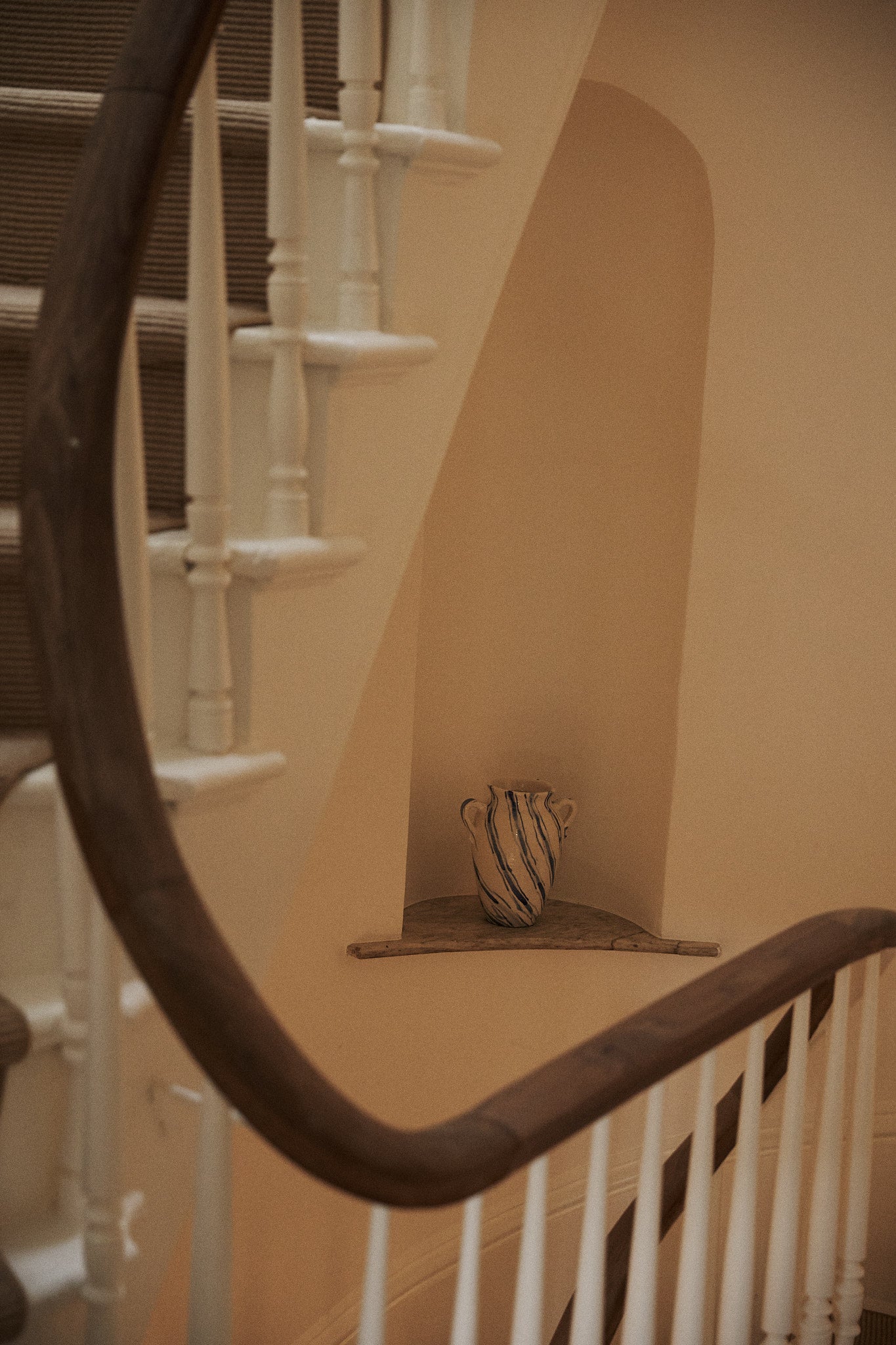 The Ida Vase, featuring blue stripes, is displayed on a small ledge beneath a curved staircase with white spindles and a wooden handrail.