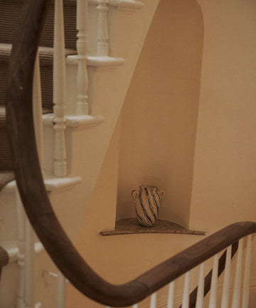 The Ida Vase, featuring blue stripes, is displayed on a small ledge beneath a curved staircase with white spindles and a wooden handrail.