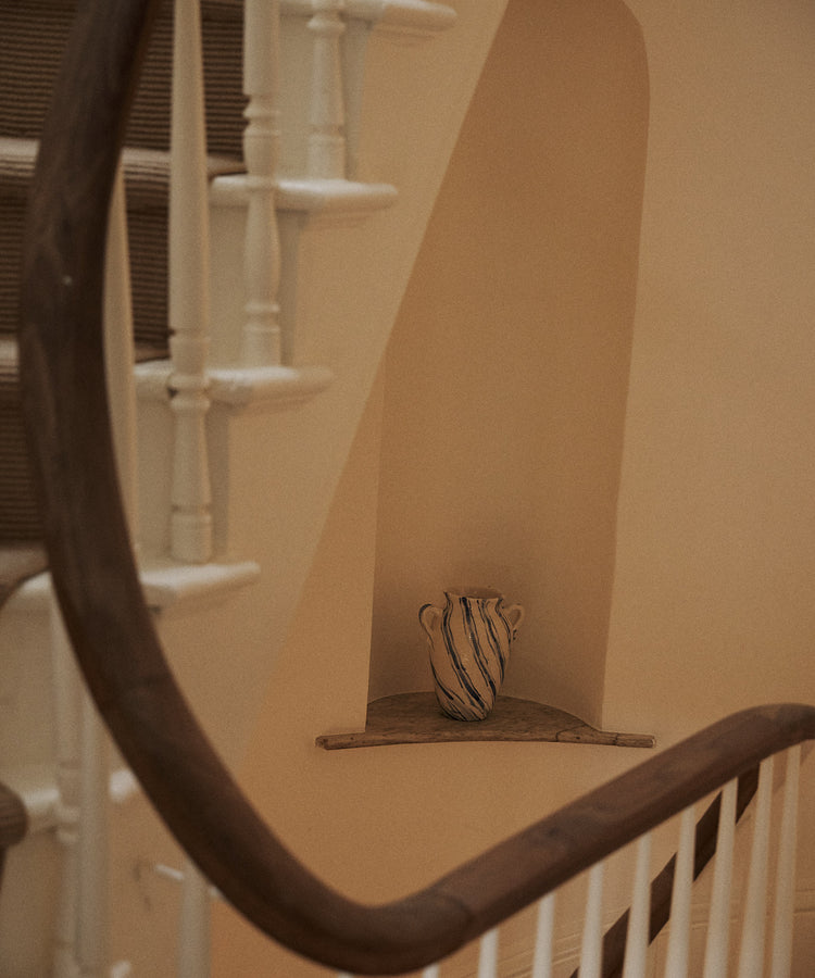 The Ida Vase, featuring blue stripes, is displayed on a small ledge beneath a curved staircase with white spindles and a wooden handrail.
