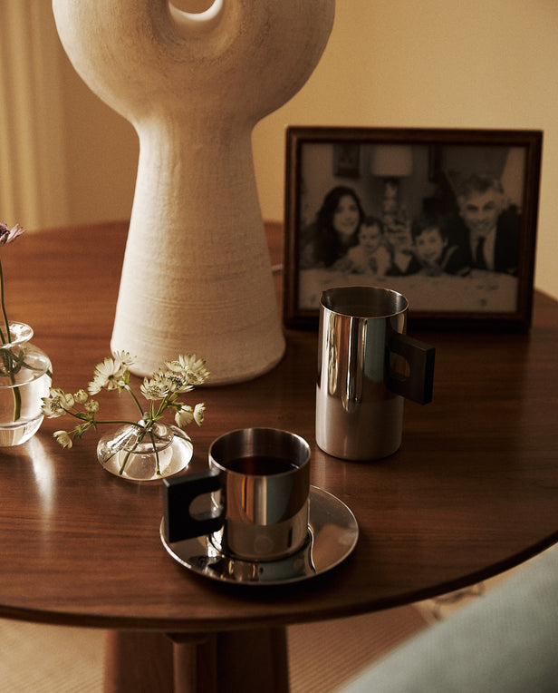 Ceramic vase, coffee cup, and silver pitcher on a wooden table with a family photo in the background.