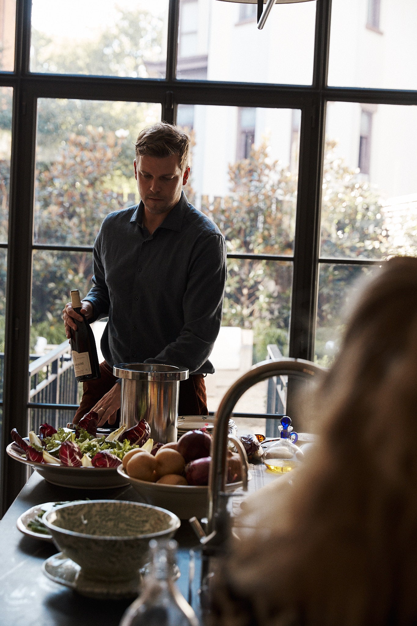A man stands at a kitchen counter holding a wine bottle next to the Metal Wine Cooler, with fruit, salad, kitchenware, and large windows in the background.
