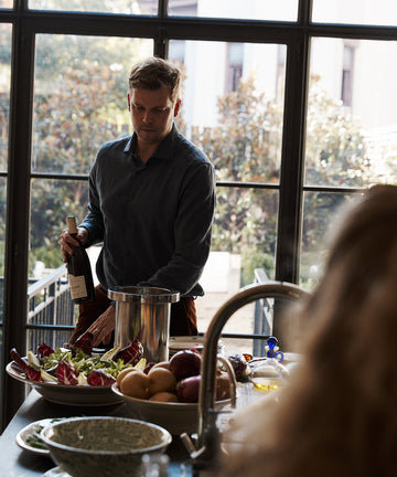 A man stands at a kitchen counter holding a wine bottle next to the Metal Wine Cooler, with fruit, salad, kitchenware, and large windows in the background.