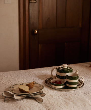 A teapot, creamer, cup, and a Betty Dip Dish filled with strawberries sit on a tray beside sliced bread and a napkin on a white tablecloth by the wooden door.