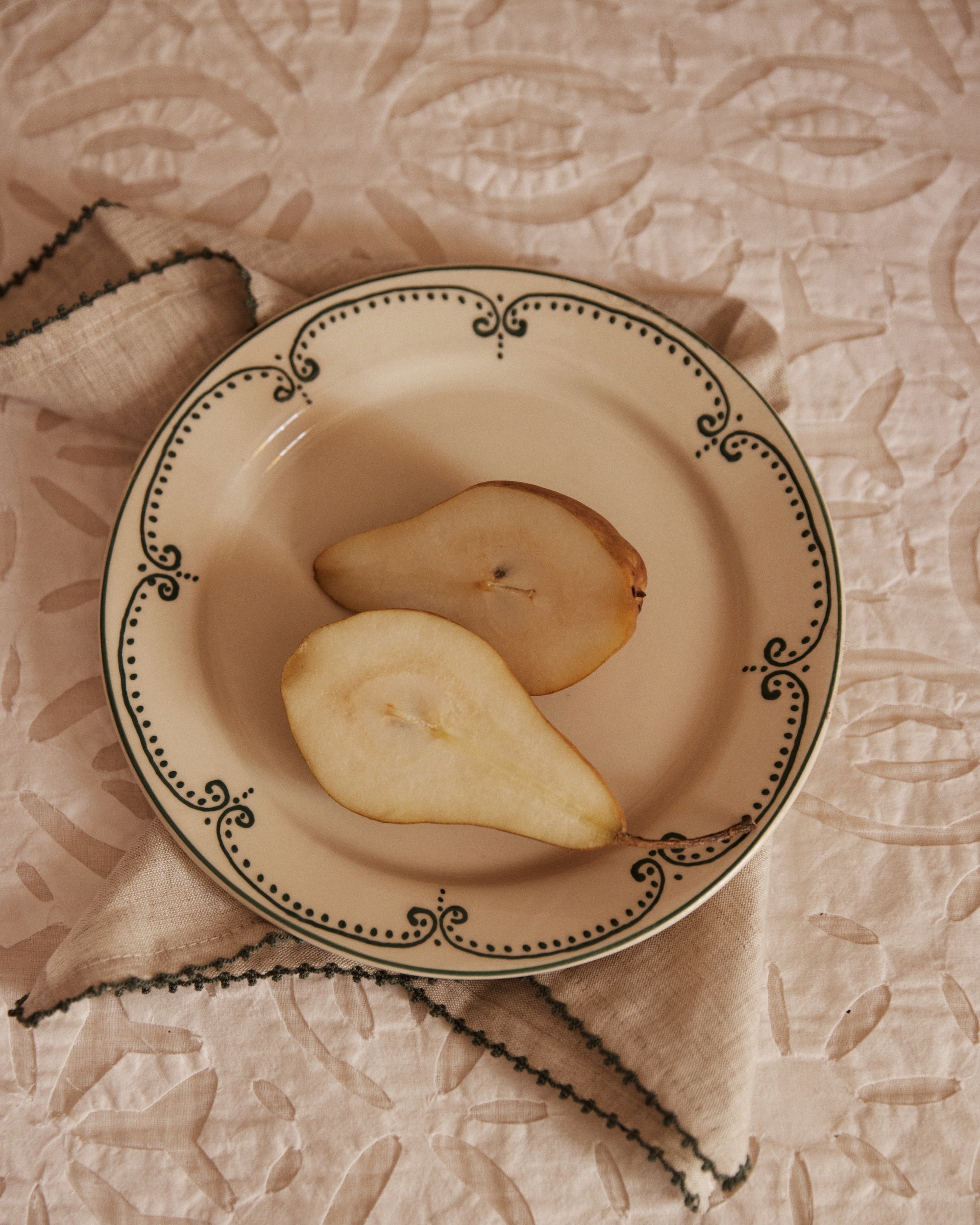 A pear sliced in half rests on the Arco Small Plate, Set of 4, with a beige napkin underneath atop a patterned tablecloth.