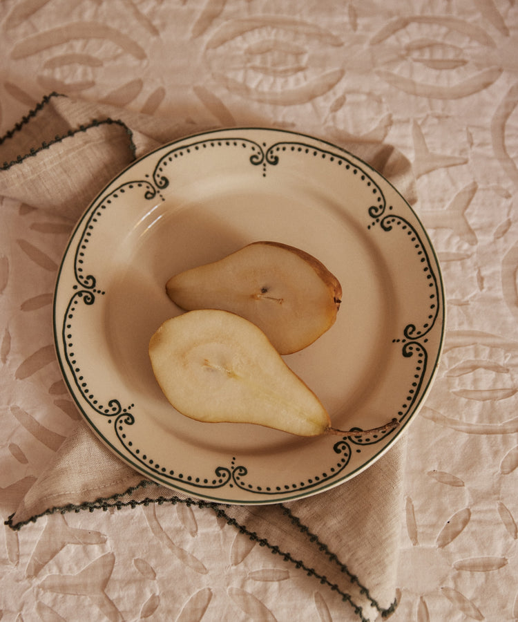 A pear sliced in half rests on the Arco Small Plate, Set of 4, with a beige napkin underneath atop a patterned tablecloth.