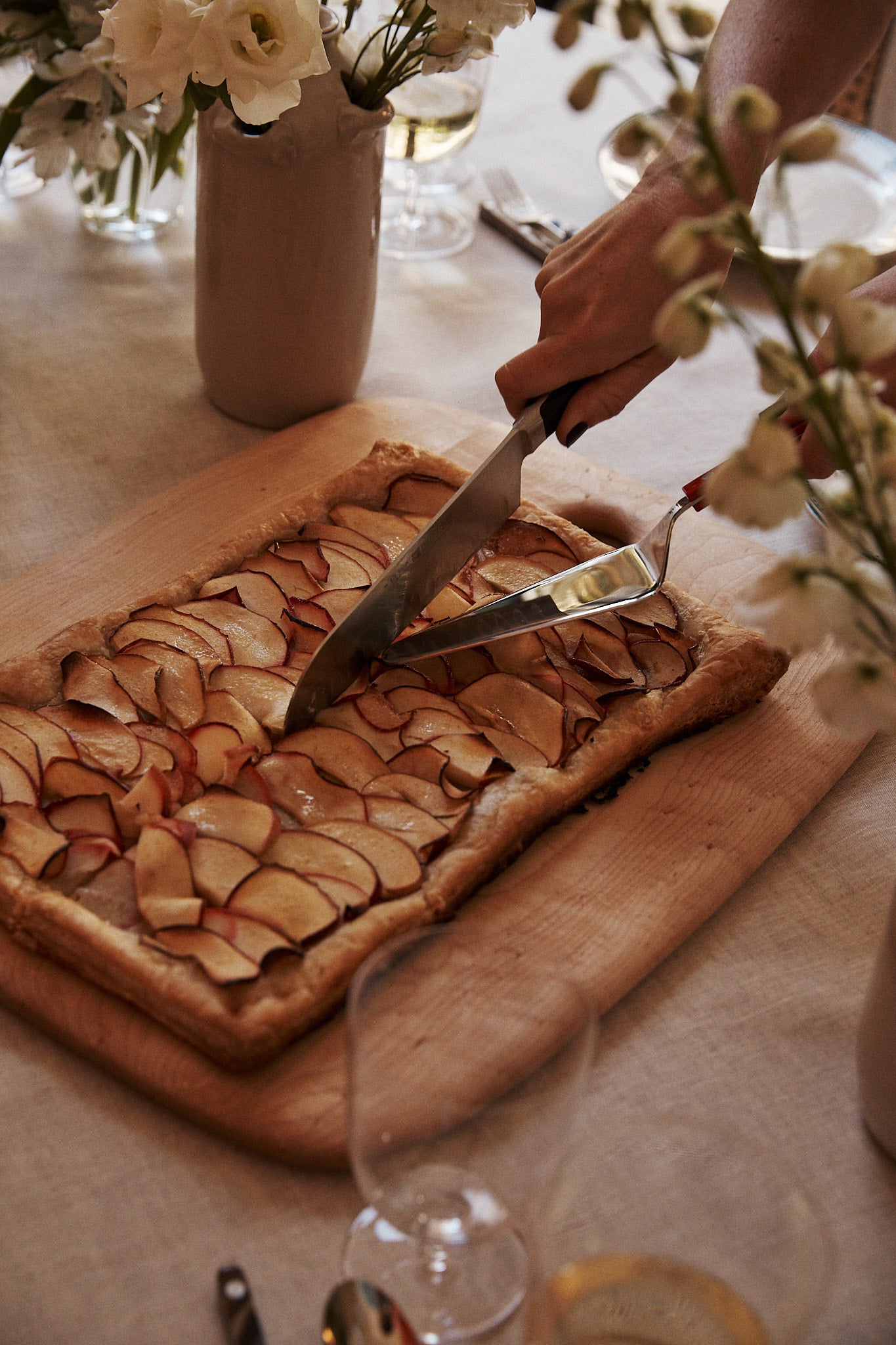 A hand cuts a rectangular fruit tart topped with thinly sliced apples on the Birds Eye Maple Chopping Board, surrounded by flowers and glassware on a table.