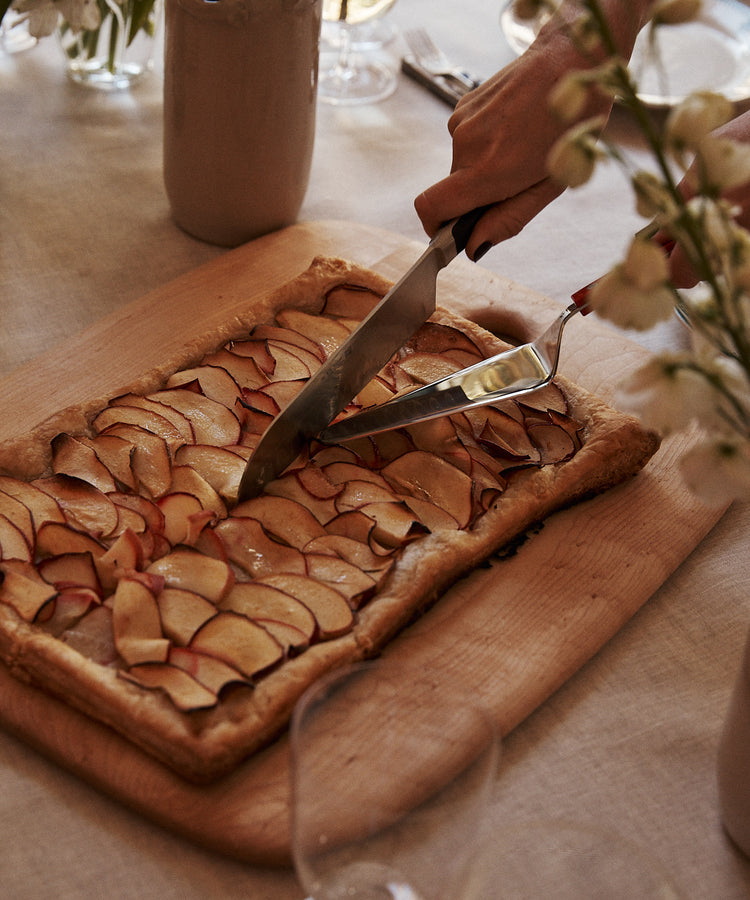 A hand cuts a rectangular fruit tart topped with thinly sliced apples on the Birds Eye Maple Chopping Board, surrounded by flowers and glassware on a table.