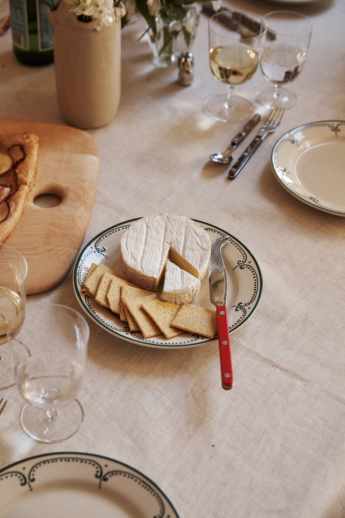A plate from the Arco Small Plate, Set of 4 displays a round wheel of soft cheese and sliced cheese, accompanied by a red-handled knife and a glass of white wine, all elegantly arranged on a set dining table.
