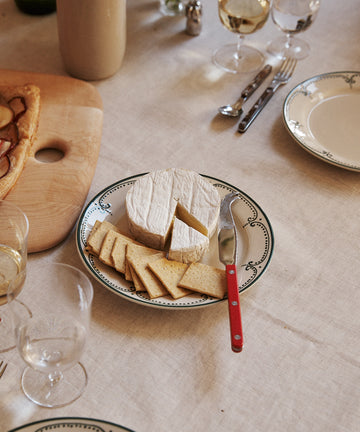 A plate from the Arco Small Plate, Set of 4 displays a round wheel of soft cheese and sliced cheese, accompanied by a red-handled knife and a glass of white wine, all elegantly arranged on a set dining table.