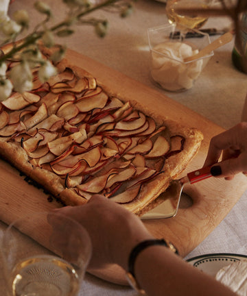A person uses a Tart Server to slice a rectangular apple tart on a wooden board, with drinks, flowers, and a cup of cream visible on the table.
