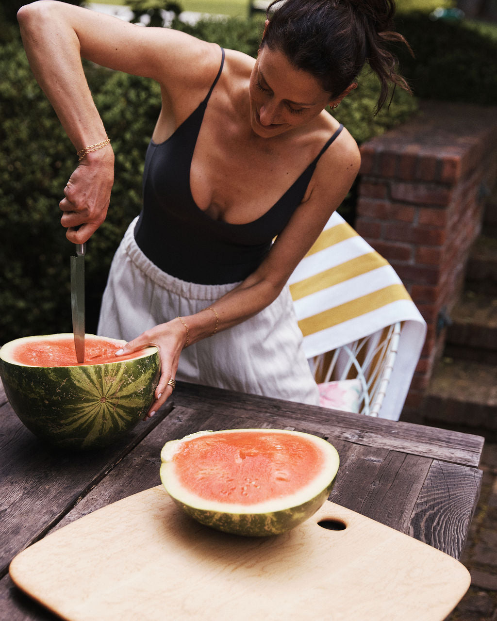 A woman slices a watermelon with a knife on a Birds Eye Maple Serving Board at an outdoor wooden table, with a striped towel and chair in the background.