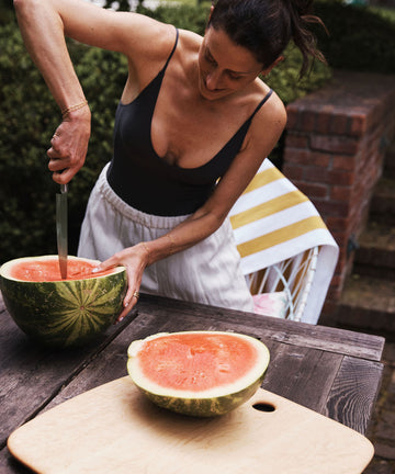 A woman slices a watermelon with a knife on a Birds Eye Maple Serving Board at an outdoor wooden table, with a striped towel and chair in the background.
