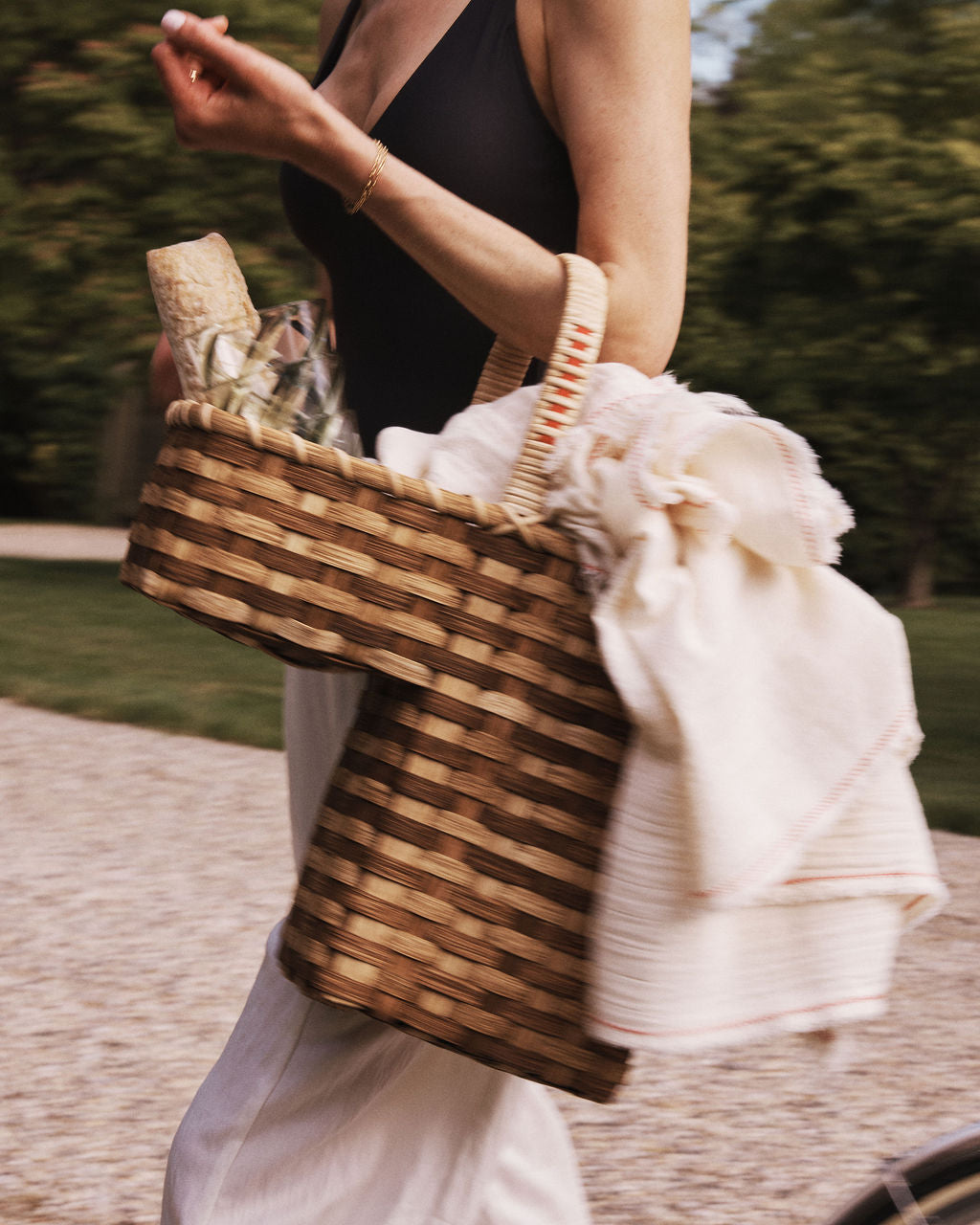 A person walks outdoors carrying the Stair Tote, a handwoven rattan basket, filled with a bottle, glassware, and a white cloth.
