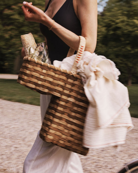 A person walks outdoors carrying the Stair Tote, a handwoven rattan basket, filled with a bottle, glassware, and a white cloth.