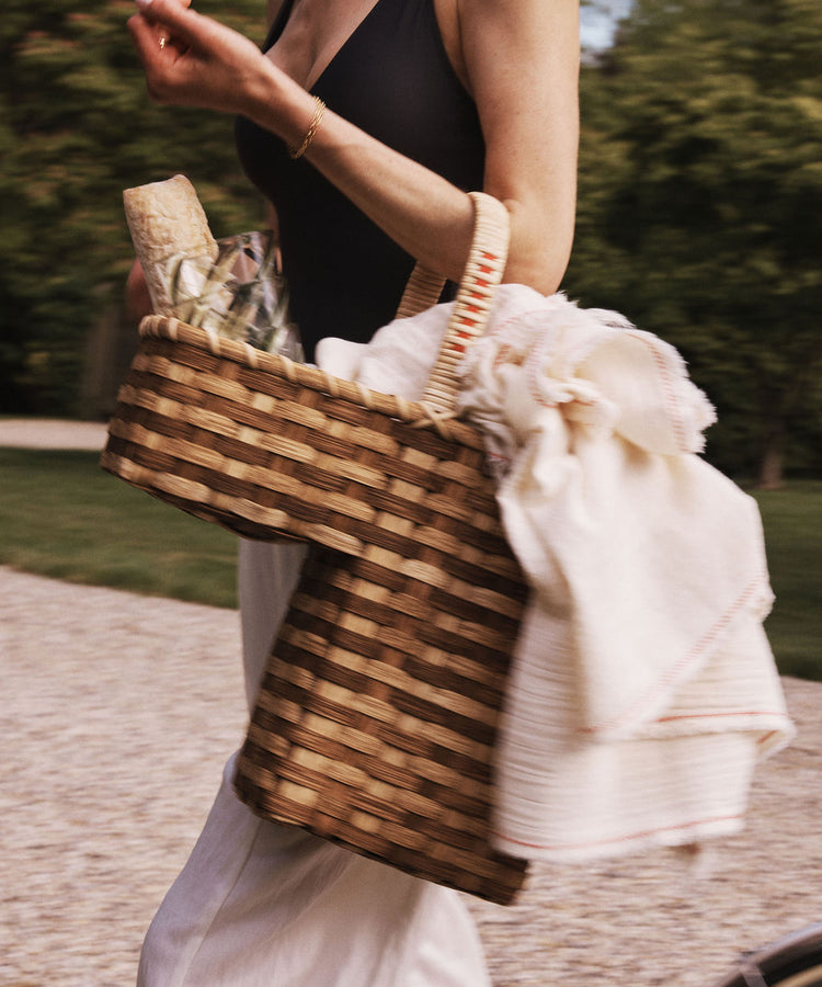 A person walks outdoors carrying the Stair Tote, a handwoven rattan basket, filled with a bottle, glassware, and a white cloth.