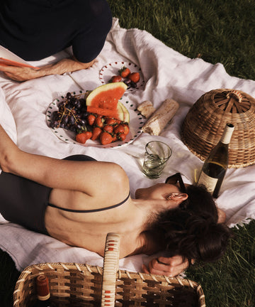 A woman relaxes on a picnic blanket with fruit, bread, wine, and baskets nearby, elegantly using the Folk Fleur Cloche as a stylish rattan food cover for outdoor entertaining.