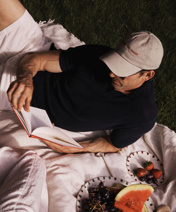 Wearing the Closed for August Cap and a dark shirt, a person relaxes on a picnic blanket, surrounded by watermelon, grapes, and strawberries, enjoying an out-of-office read.