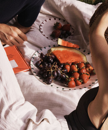 Watermelon slices, grapes, and strawberries are arranged on the Nina Serving Plate atop a picnic blanket, with two people sitting nearby and a red book beside them.