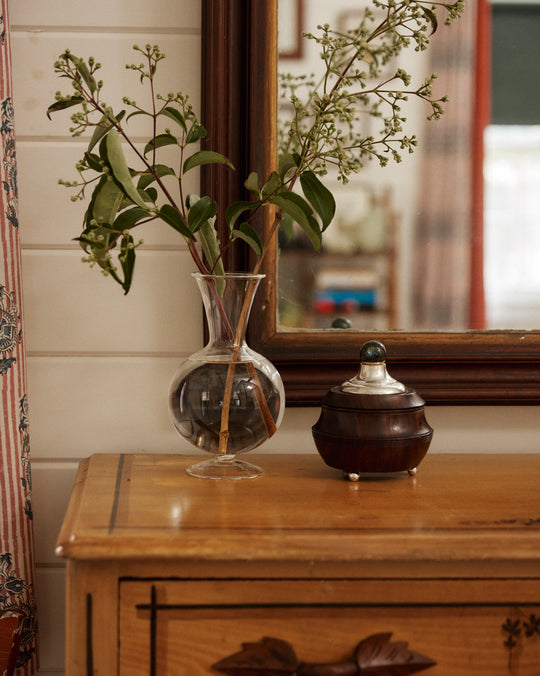The Boro Vase with green branches sits alongside a round wooden container on a wooden dresser in front of a mirror.