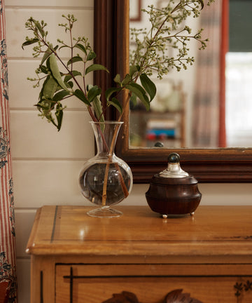 The Boro Vase with green branches sits alongside a round wooden container on a wooden dresser in front of a mirror.