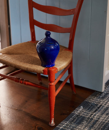 The Hugo Money Pot sits on a woven-seat red chair, with a blue door and patterned rug in the background.