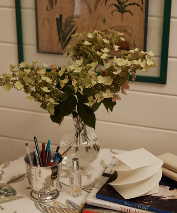 A Boro Vase of hydrangeas sits on a table with pens in a cup, a perfume bottle, stacked books, notepaper, and a botanical print on the wall behind.