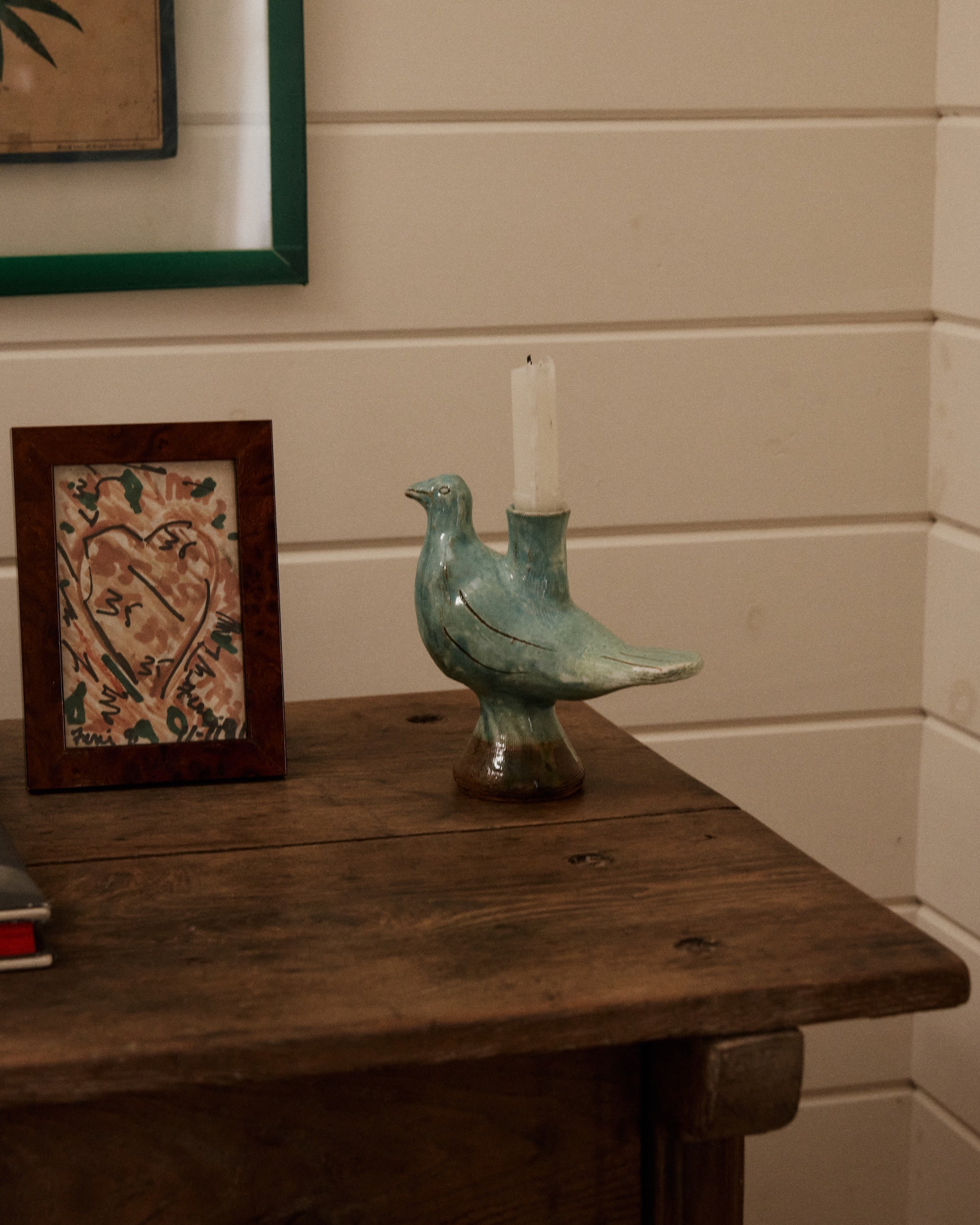 The Peace Dove Candle Holder with a sage green glaze holds a white candle on a wooden table beside a framed heart drawing against a white paneled wall.