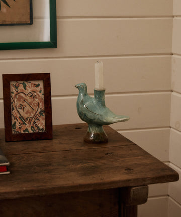 The Peace Dove Candle Holder with a sage green glaze holds a white candle on a wooden table beside a framed heart drawing against a white paneled wall.