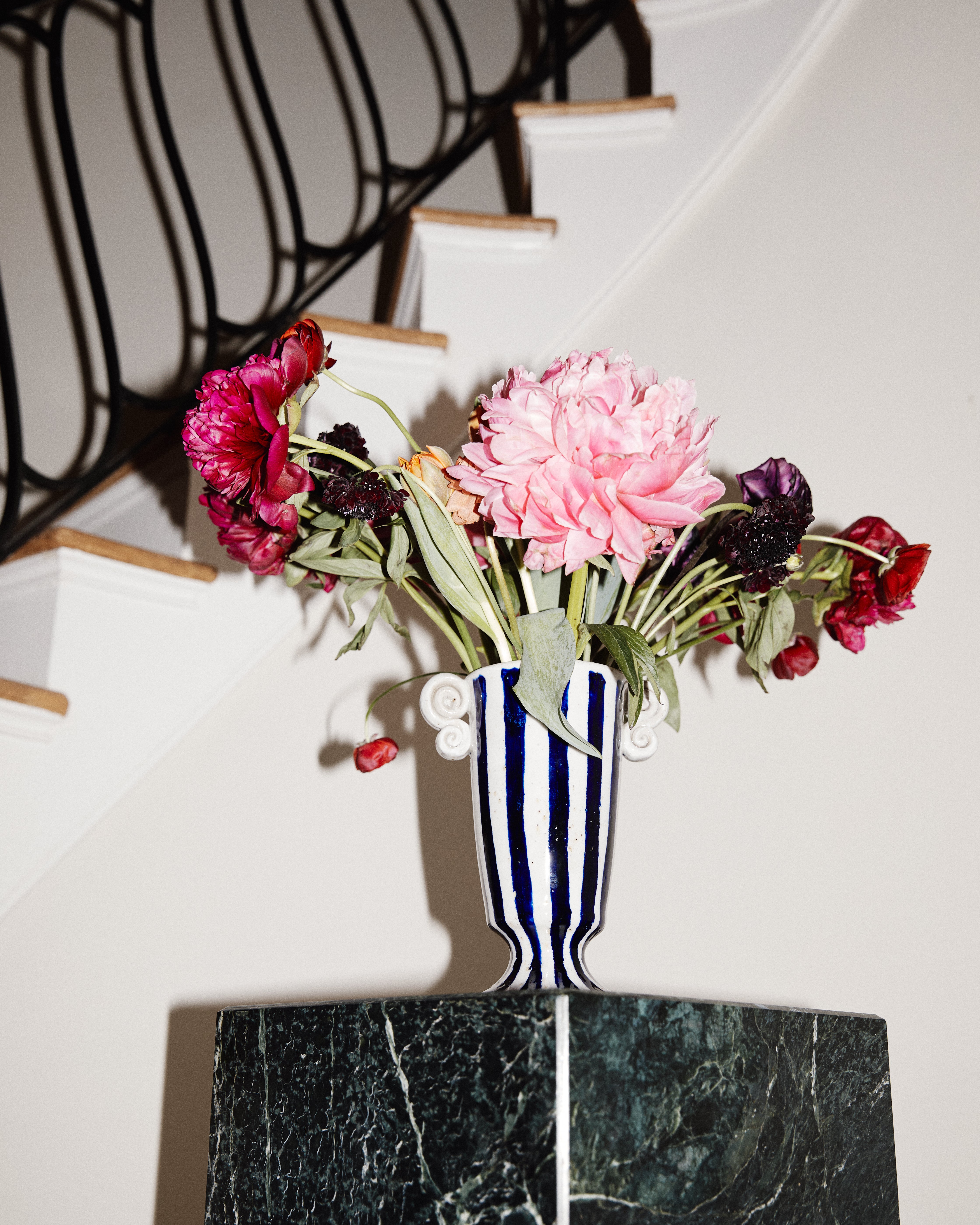 The Thallo Pedestal Vase, hand-painted in blue and white and filled with wilted pink and red flowers, stands on a green marble pedestal before a black-railed staircase, making it a striking statement piece.