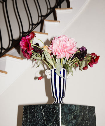The Thallo Pedestal Vase, hand-painted in blue and white and filled with wilted pink and red flowers, stands on a green marble pedestal before a black-railed staircase, making it a striking statement piece.